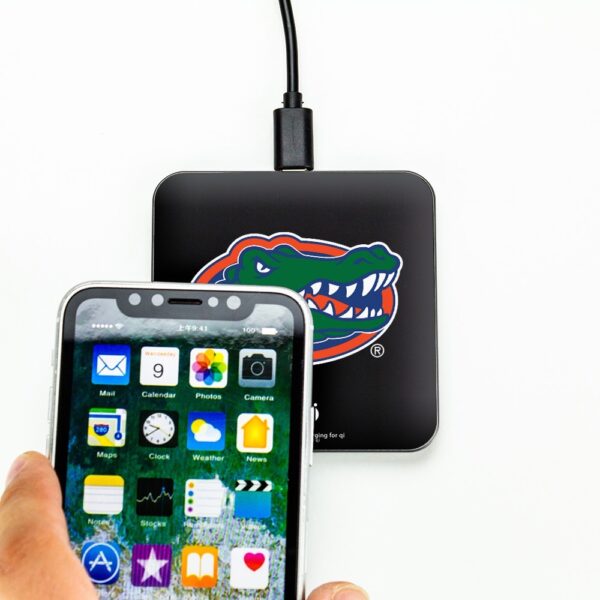 A hand holds a smartphone with app icons displayed, connected via cable to a Florida Gators QuikCharge Wireless Charger - Qi Certified.