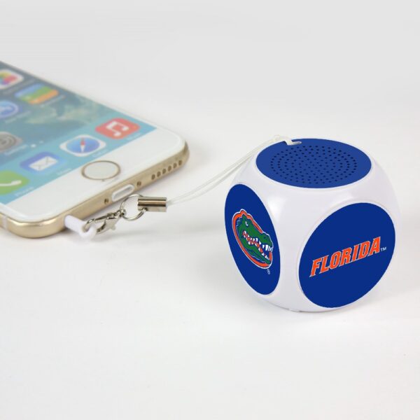 A small, blue and white portable speaker labeled "Florida Gators MX-100 Cubio Mini Bluetooth® Speaker Plus Selfie Remote" connected to a smartphone via an audio cable, placed on a white background.