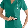 A smiling healthcare professional in a CROSS FLEX V-NECK MEDIA SCRUB TOP C12110 puts a device in her pocket, wearing a stethoscope around her neck.