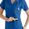 A smiling healthcare worker in a CROSS FLEX V-NECK MEDIA SCRUB TOP C12110 places a digital device into her pocket, with a stethoscope around her neck.