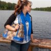 A woman with a ponytail, wearing a black shirt and blue scarf, holding The Endall Handbag (Florida Gators), stands by a lake railing, looking away thoughtfully.