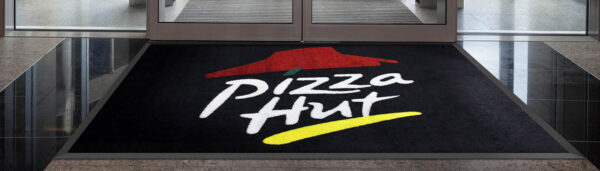 A Custom Carpet Logo Mat with the Pizza Hut emblem, featuring a red hat and the name in white letters on a black background, located at a building entrance.