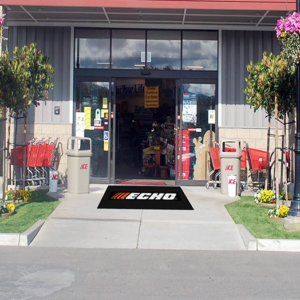 Entrance of a hardware store with an Hi Res Logo Scraper Mats welcome mat, shopping carts to the left, potted plants, and visible interior shelves stocked with items.