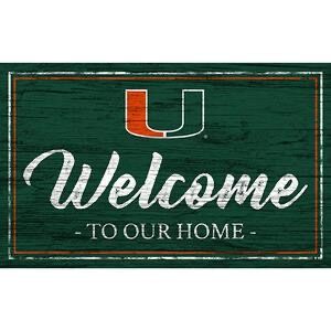 A vintage-style green wooden sign with the word "welcome" in large, white cursive letters, and "to our home" in smaller script, featuring a red horseshoe as the letter "u.
