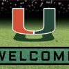 Miami Hurricanes Crumb Rubber Door Mat featuring a green and orange "u" above a "welcome" sign, set against a backdrop of a grass field with a blurred stadium in the distance.