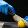 A person wearing Gloveworks Blue Vinyl Industrial Latex Free Disposable Gloves (Case of 1000) cuts a lemon on a black cutting board with a stainless steel knife.