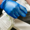 A hand in Gloveworks Blue Vinyl Industrial Latex Free Disposable Gloves holds a plastic container with a green label, examining its contents.