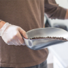 A person wearing Gloveworks Clear Vinyl Industrial Latex Free Disposable Gloves holding a metal scoop filled with coffee beans.